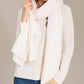 A woman stands against a plain background, her face partially visible, wearing a white sweater and the Alashan Cashmere Luxe Travel Wrap. She holds the luxurious cashmere wrap in one hand.