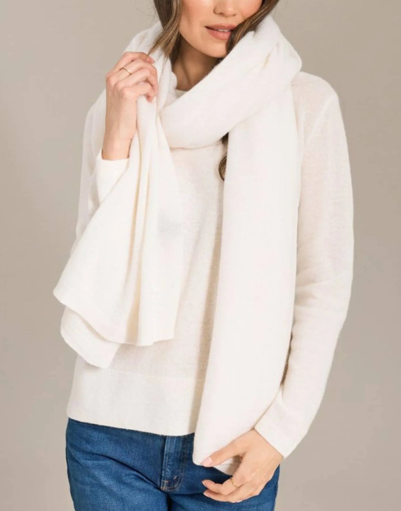 A woman stands against a plain background, her face partially visible, wearing a white sweater and the Alashan Cashmere Luxe Travel Wrap. She holds the luxurious cashmere wrap in one hand.