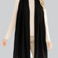 A woman wears the Alashan Cashmere Luxe Travel Wrap—draped long and black—over a cream sweater and beige pants, standing against a plain light gray background.