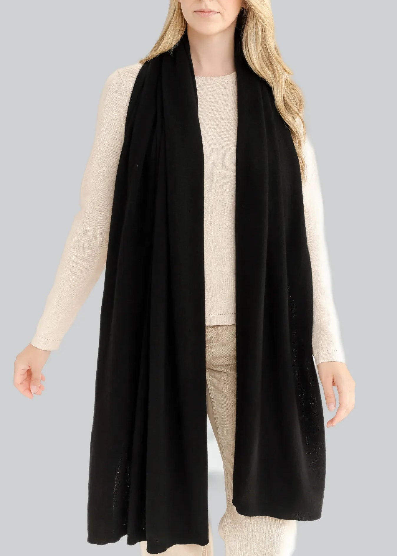 A woman wears the Alashan Cashmere Luxe Travel Wrap—draped long and black—over a cream sweater and beige pants, standing against a plain light gray background.