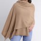 A woman wearing a light brown draped poncho-style sweater and light blue jeans, elegantly accessorized with the Alashan Cashmere Cashmere Luxe Travel Wrap, stands against a plain white background.