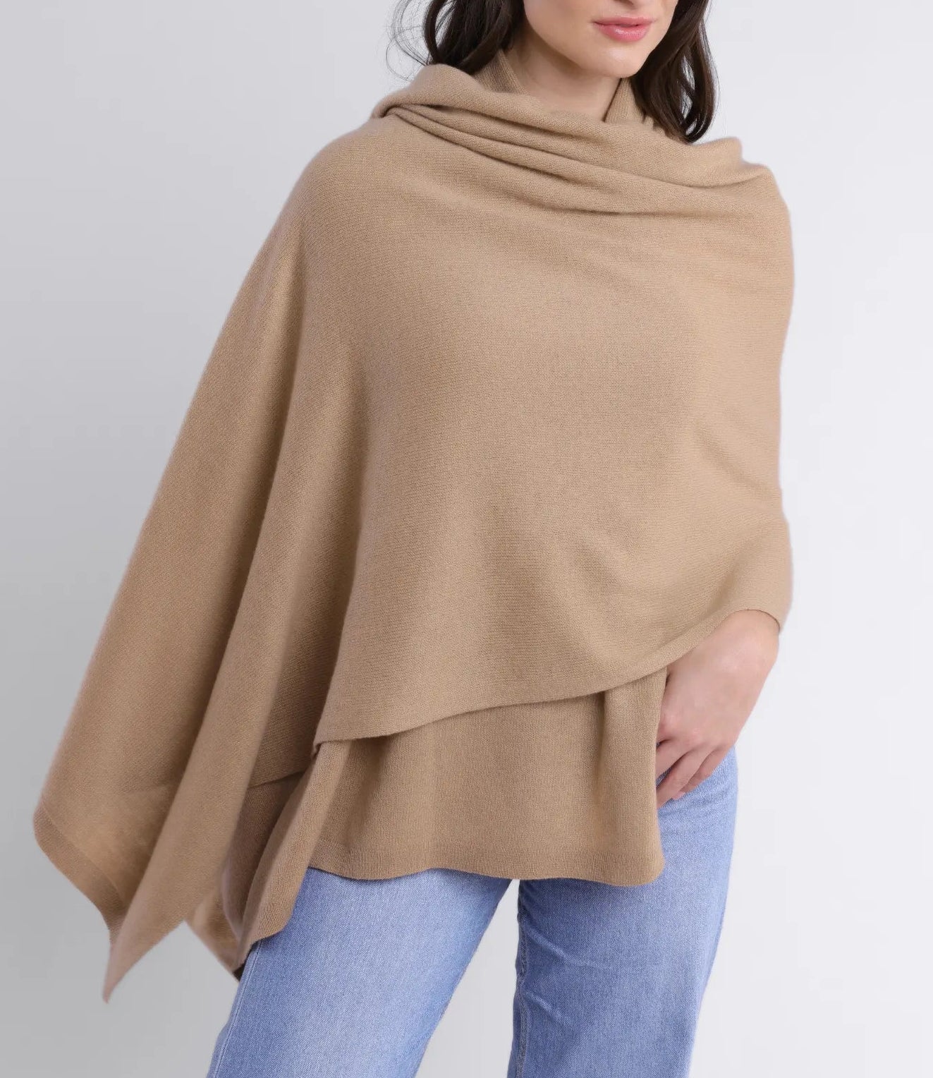 A woman wearing a light brown draped poncho-style sweater and light blue jeans, elegantly accessorized with the Alashan Cashmere Cashmere Luxe Travel Wrap, stands against a plain white background.