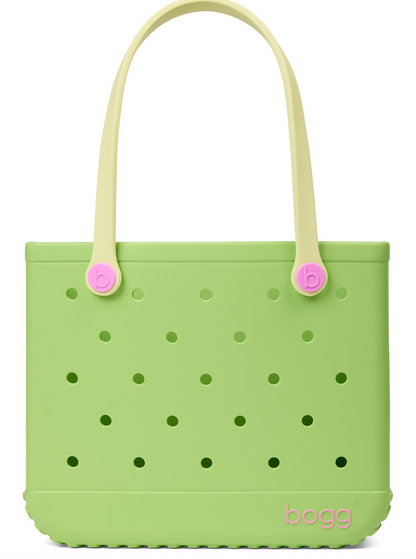A light green Baby Bogg® Bag tote from Bogg Bags, featuring evenly spaced holes, cream-colored handles with pink accents, and the word “bogg” printed in pink in the lower corner. This durable bag is fully washable for easy care and long-lasting use.