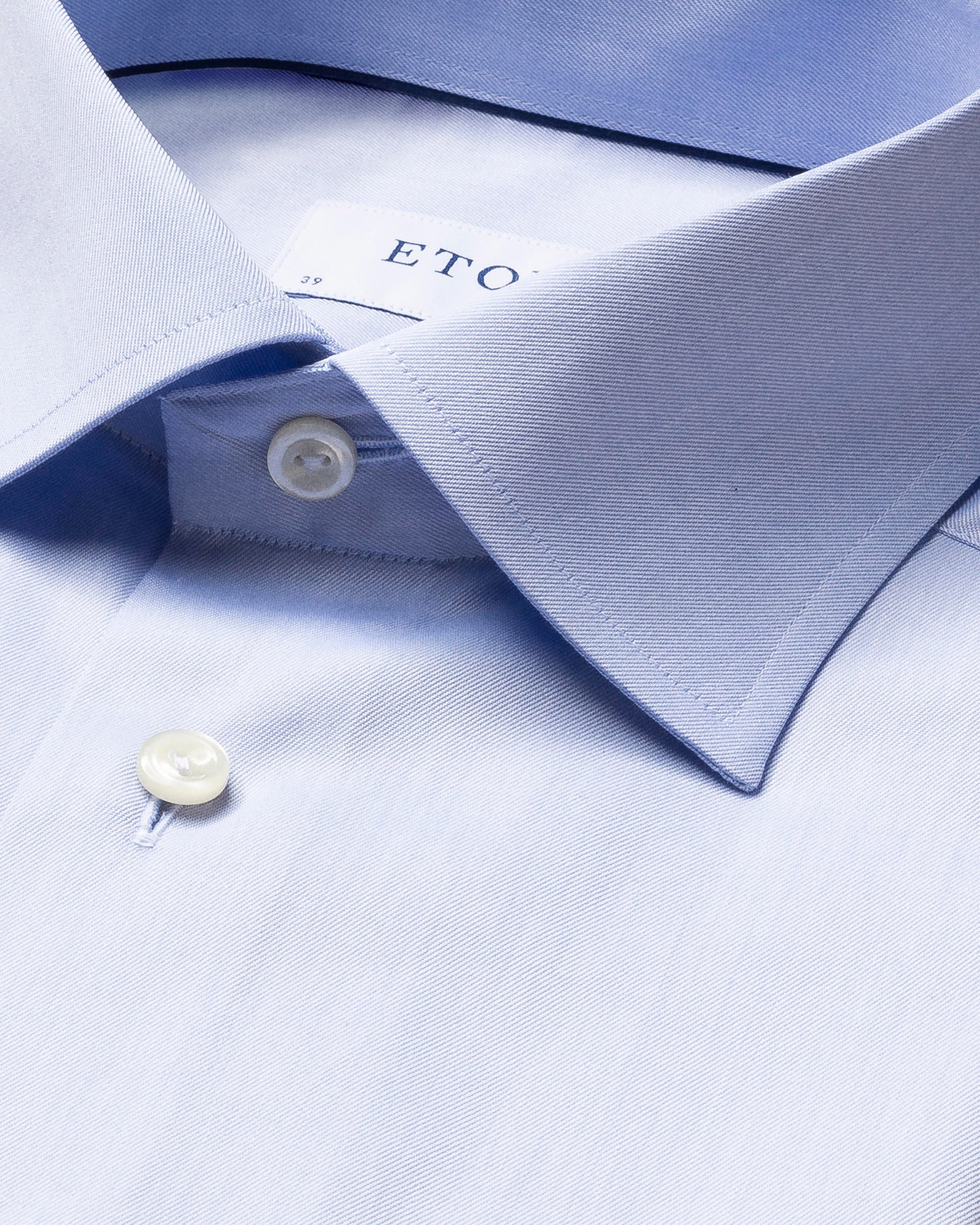 Close-up of the light blue Eton Light Blue Signature Twill Shirt in a slim fit, featuring a pointed collar and two visible buttons. The brand's signature twill fabric and wrinkle-free design are highlighted, with a partial view of the "ETON" label visible.