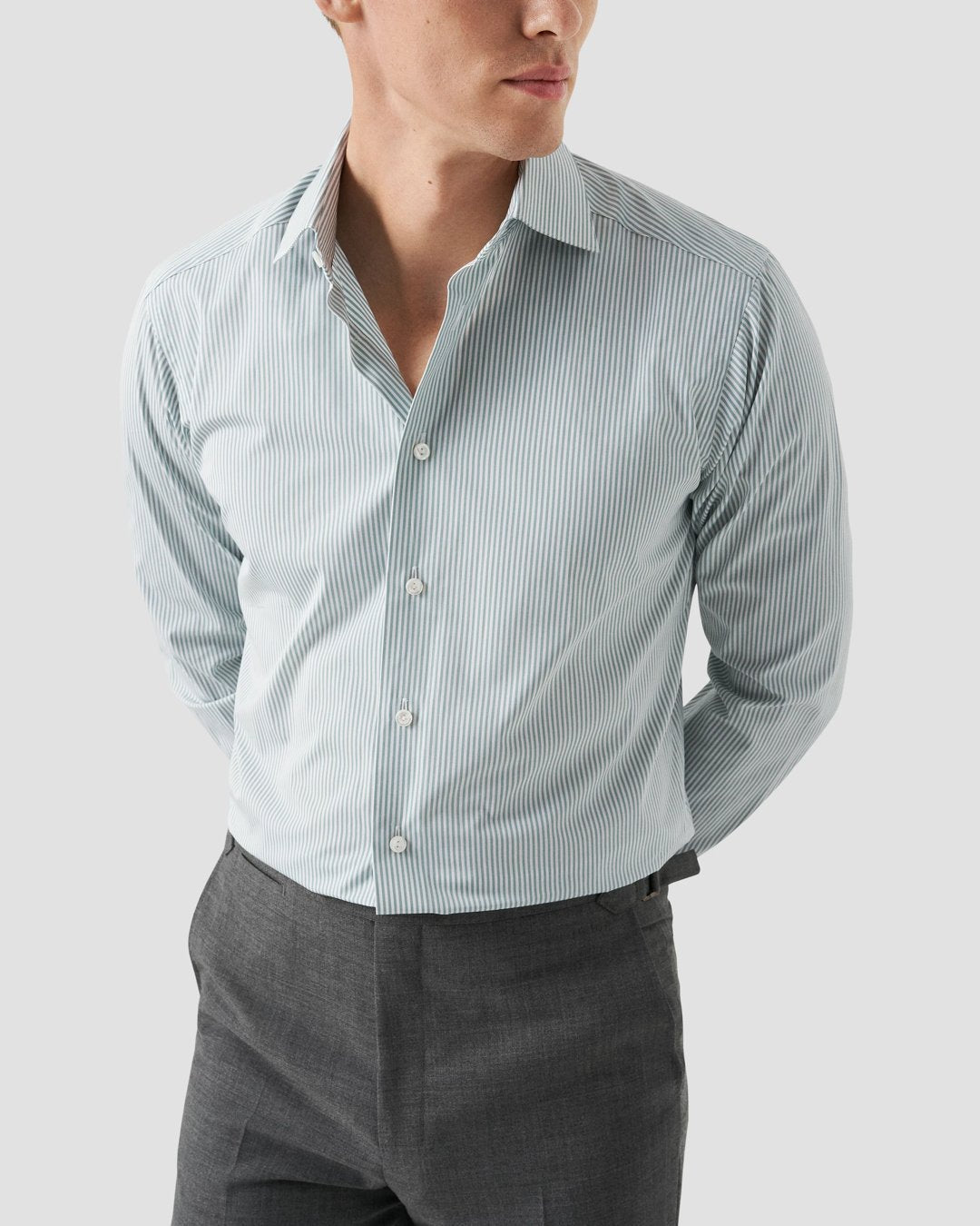 A person wearing an Eton Bengal Stripe Poplin Shirt, Slim Fit in light blue, paired with grey trousers, has their hands behind their back and is looking slightly to the side.