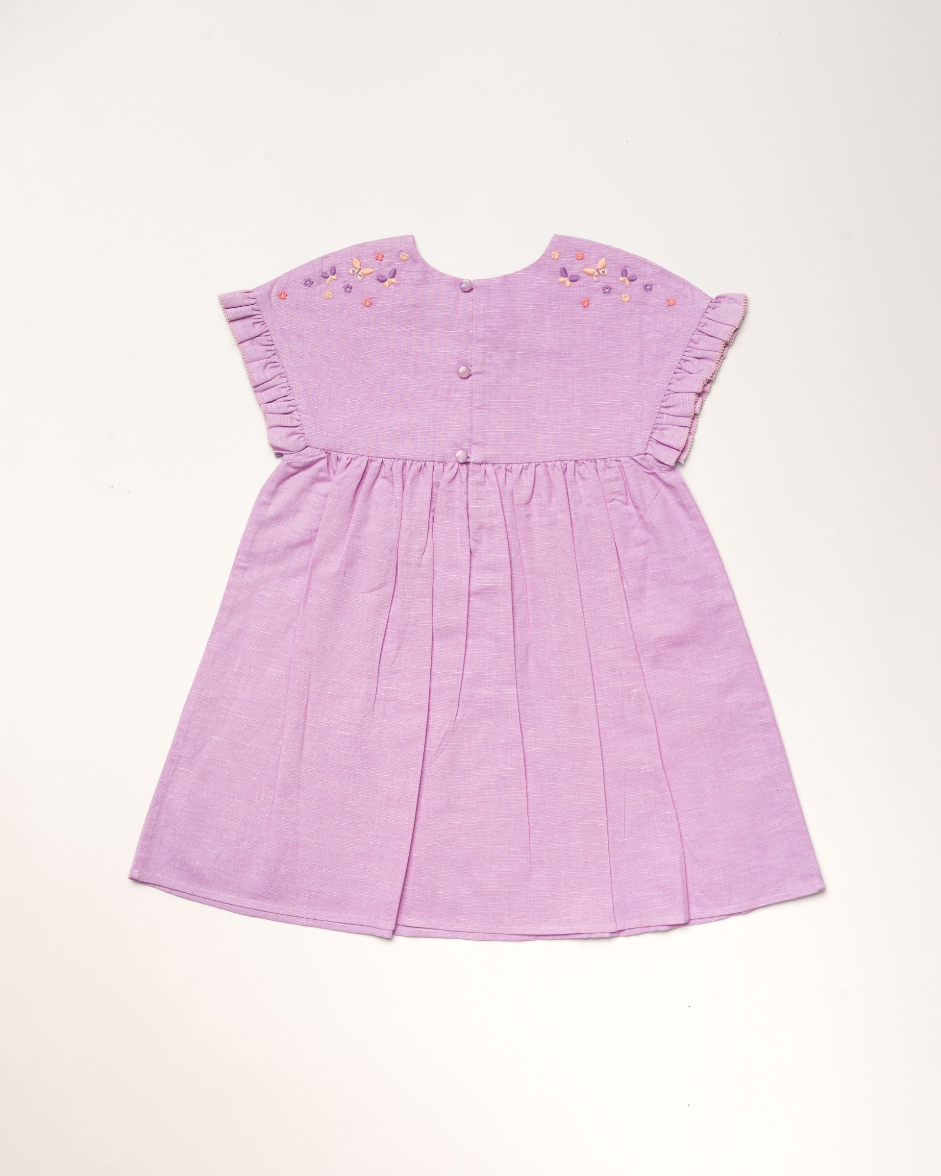 The Early Sunday Girls’ Lila Dress is a short-sleeved lilac dress featuring a pleated skirt, flutter sleeves, embroidered flowers on the shoulders, and back buttons, displayed against a light background.