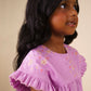 A young girl with long dark hair, wearing the Early Sunday Girls’ Lila Dress—a charming cotton-linen piece in lilac with embroidered flowers and flutter sleeves—looks upward and smiles against a plain beige background.