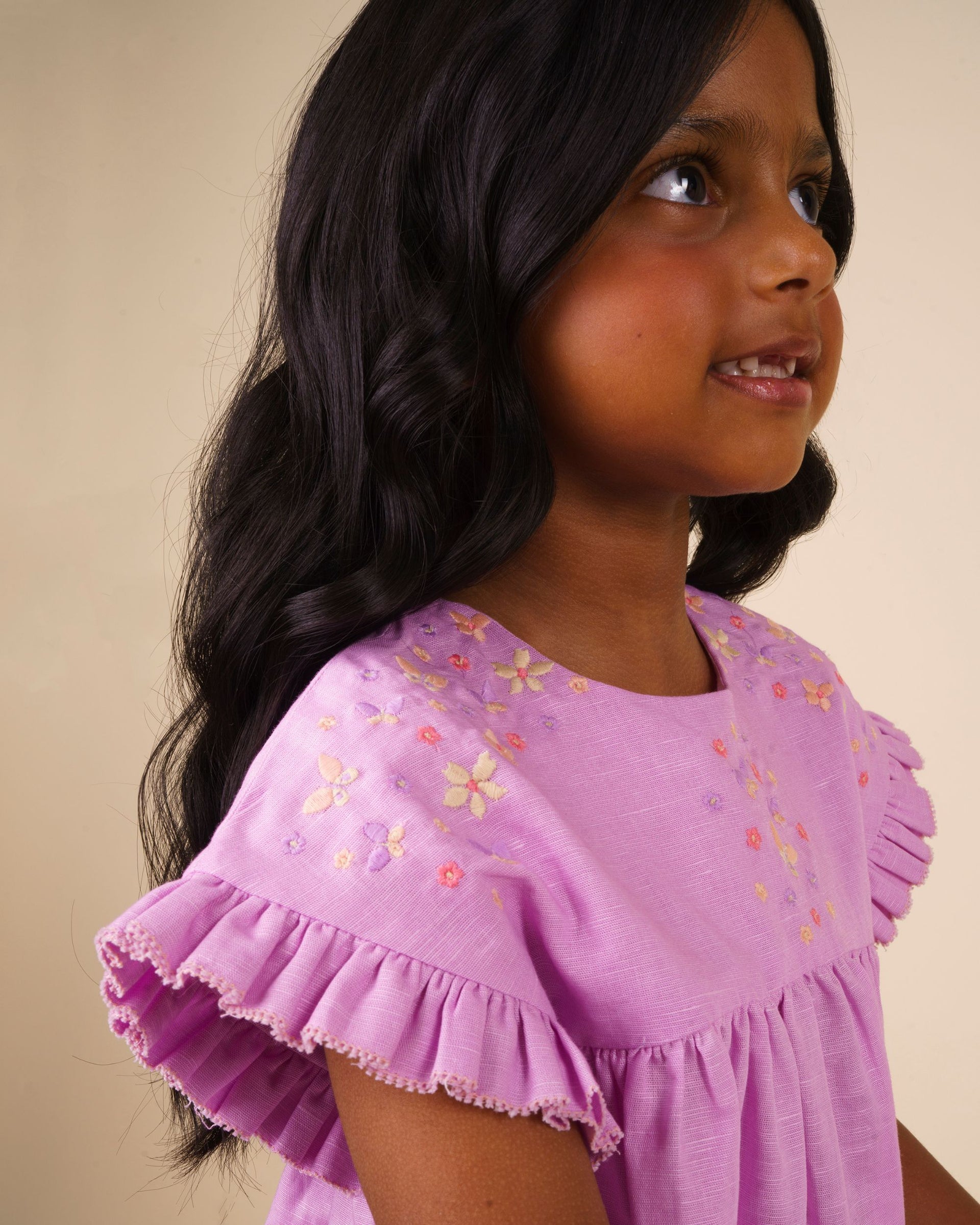 A young girl with long dark hair, wearing the Early Sunday Girls’ Lila Dress—a charming cotton-linen piece in lilac with embroidered flowers and flutter sleeves—looks upward and smiles against a plain beige background.
