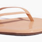 Close-up of Tkees Lily Glosses Sandals in beige patent leather with a glossy finish, featuring a toe strap and part of the sole crafted from Brazilian leather, shown against a white background.