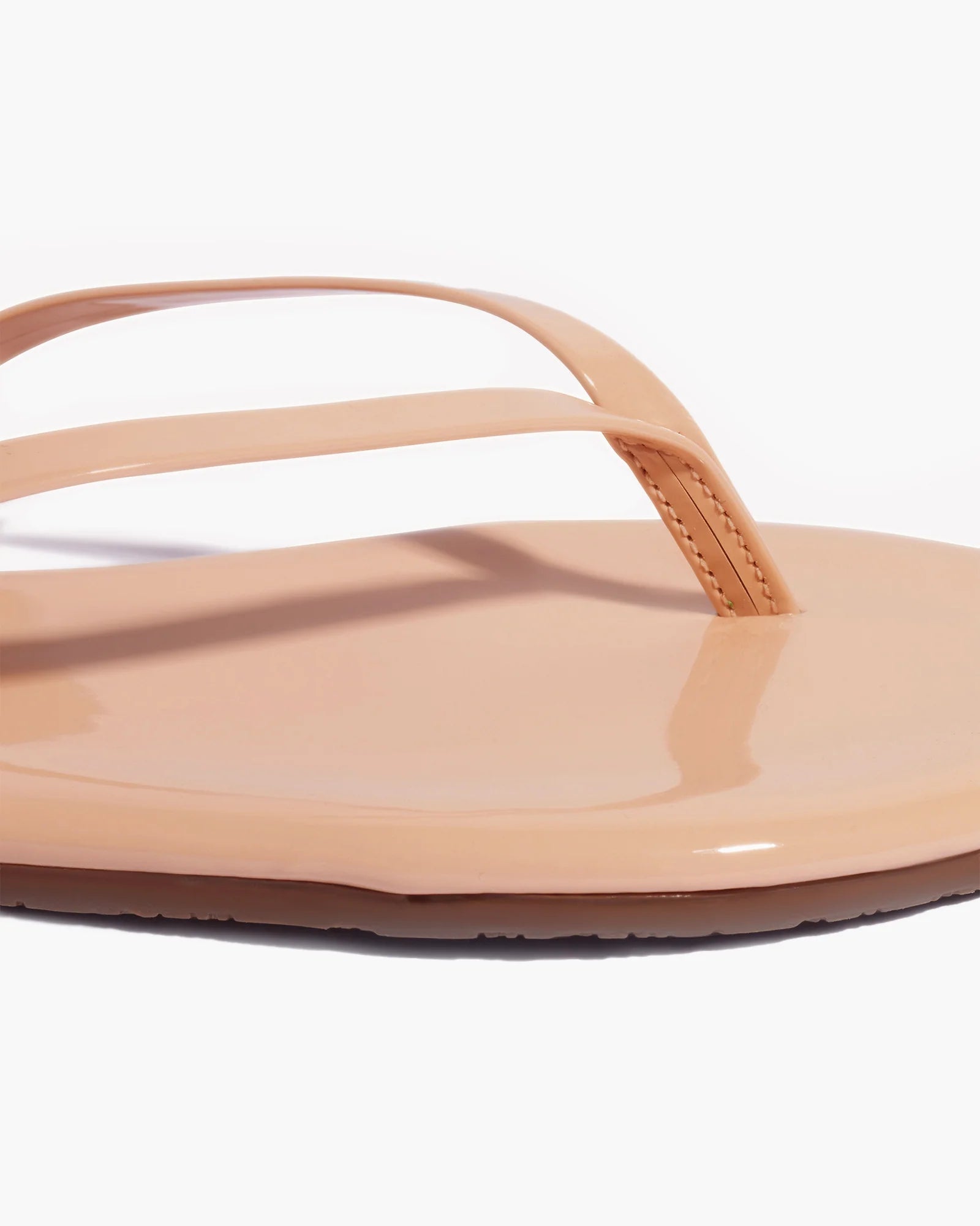 Close-up of Tkees Lily Glosses Sandals in beige patent leather with a glossy finish, featuring a toe strap and part of the sole crafted from Brazilian leather, shown against a white background.