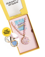 The Super Smalls Sparkle Shell Lipgloss Locket is a pink and pearl necklace with a shell-shaped pendant filled with coconut-scented gloss, displayed in an open yellow “Lip Gloss Lockets” box.