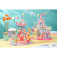 Two colorful Calico Critters playsets, including the Baby Mermaid Castle, with small animal baby figures are displayed on a table with a pastel background, flowers, jars, bunting, and an open book in the foreground. The Baby Mermaid Castle playset features a delightful sea-themed amusement park and mermaid castle for endless imaginative adventures.