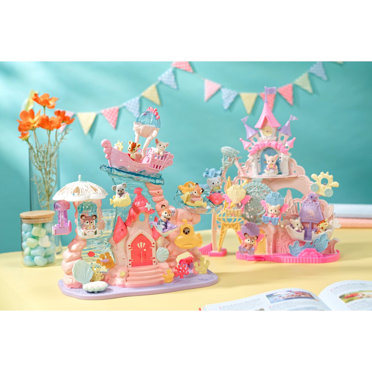Two colorful Calico Critters playsets, including the Baby Mermaid Castle, with small animal baby figures are displayed on a table with a pastel background, flowers, jars, bunting, and an open book in the foreground. The Baby Mermaid Castle playset features a delightful sea-themed amusement park and mermaid castle for endless imaginative adventures.
