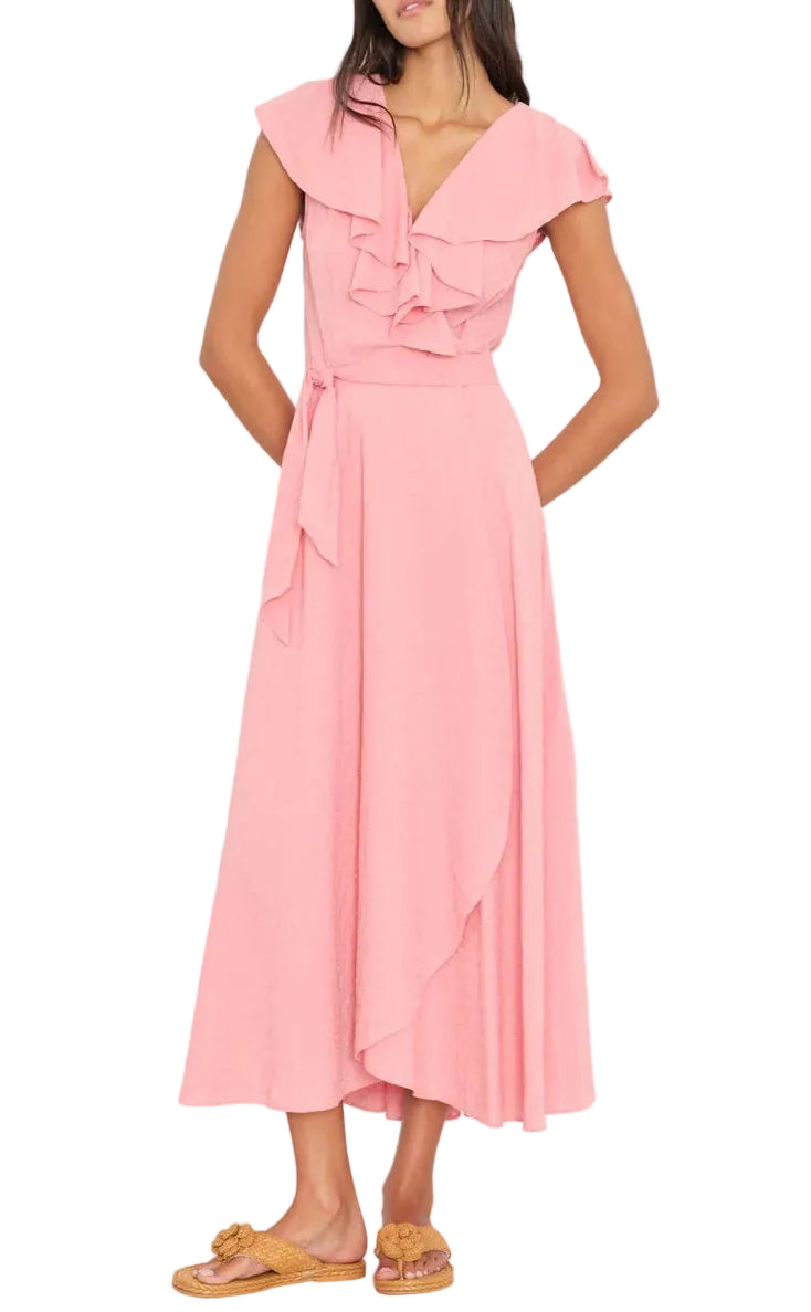 A woman wears the Xirena Lola Dress, a sleeveless light pink wrap dress with ruffle details and a waist tie in soft cotton poplin, paired with tan sandals.