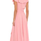 A woman stands facing away, wearing the Xirena Lola Dress—a pink cotton poplin ankle-length dress with ruffled sleeves and a belt—paired with tan flat sandals.
