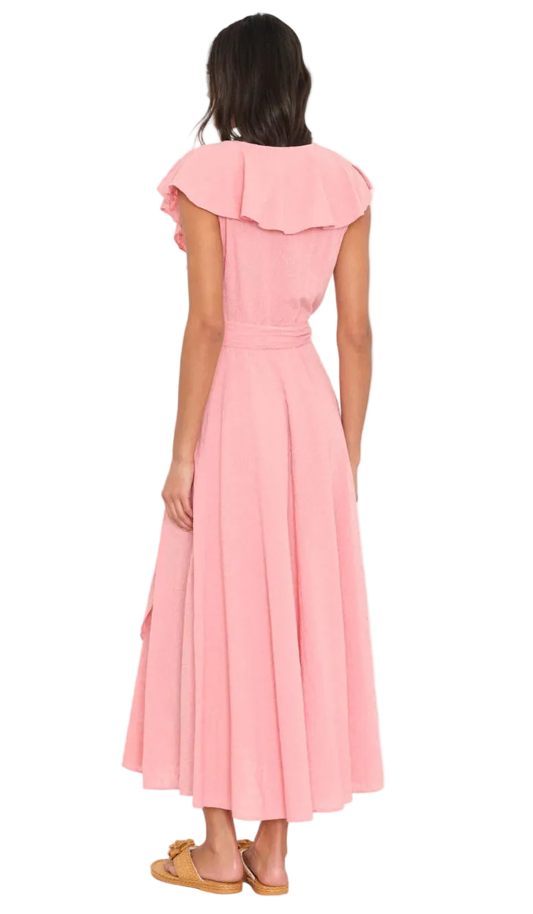 A woman stands facing away, wearing the Xirena Lola Dress—a pink cotton poplin ankle-length dress with ruffled sleeves and a belt—paired with tan flat sandals.