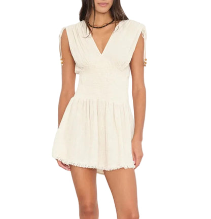 A woman wears the Xirena Thelma Dress, a cream-colored bohemian mini with a V-neckline, smocked waist, and gathered skirt, standing against a white background.