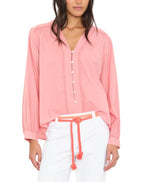 A woman wears the Xirena Dany Shirt, an airy pink blouse with lace inset detailing, paired with white pants and a coral rope belt, standing against a plain white background.