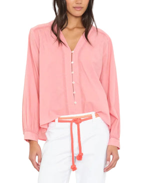 A woman wears the Xirena Dany Shirt, an airy pink blouse with lace inset detailing, paired with white pants and a coral rope belt, standing against a plain white background.