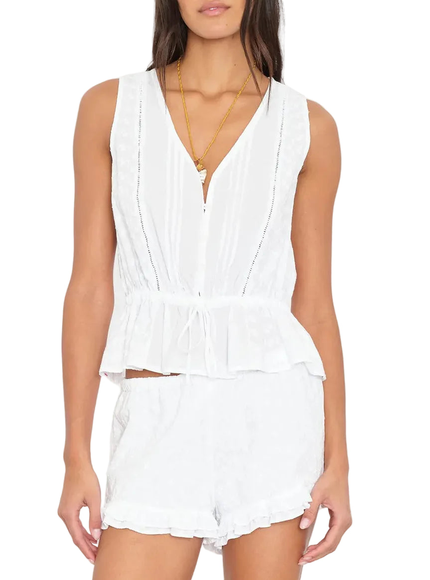 A woman wears the Emma Top by Xirena, a sleeveless floral-embroidered peplum piece, paired with matching white shorts and a gold necklace.