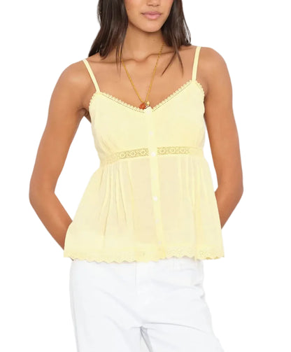 A woman wears the Xirena Alexia Top—a sleeveless yellow camisole with lace trim—paired with white pants, standing with her hands in her pockets.