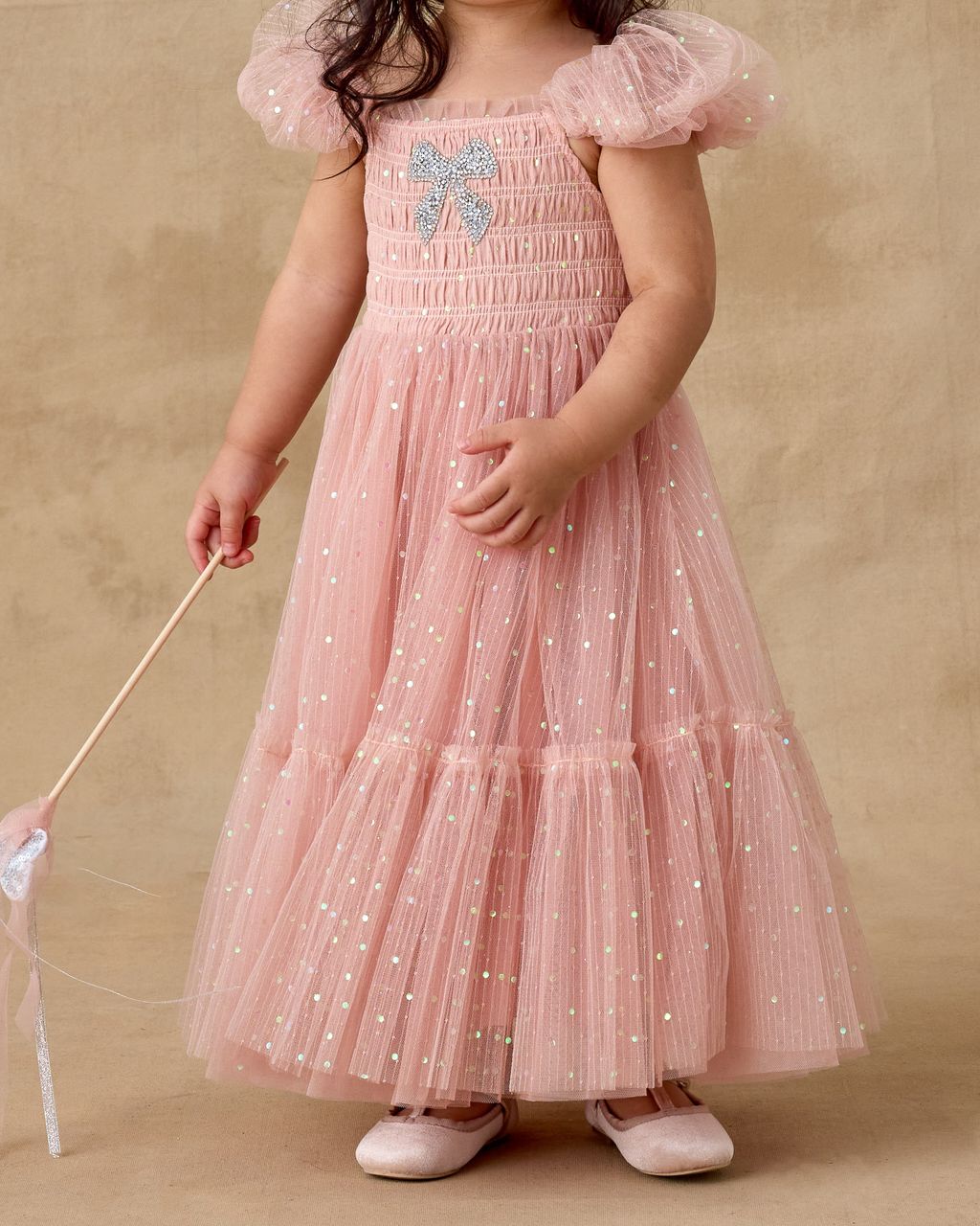 A young child in the Noralee Clara Dress—a pink, glittery dress with smocked bodice, puffed sleeves, and bow detail—holds a wand while standing against a beige background.