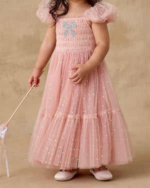 A young child in the Noralee Clara Dress—a pink, glittery dress with smocked bodice, puffed sleeves, and bow detail—holds a wand while standing against a beige background.