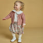 A young child with curly blond hair wears the Kidiwi Baby Girls' Lorena Blouse with a Peter Pan collar over a floral dress, paired with white tights and shoes, standing against a plain beige background.