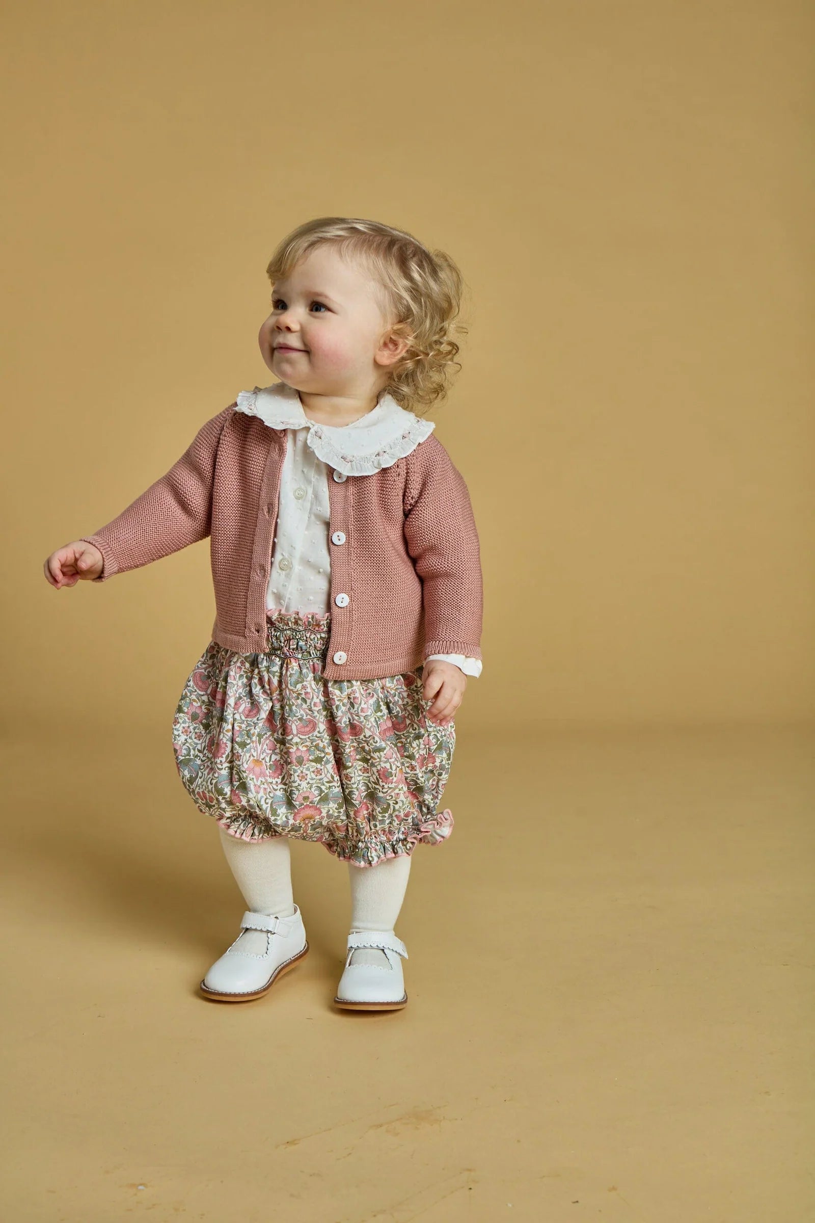 A young child with curly blond hair wears the Kidiwi Baby Girls' Lorena Blouse with a Peter Pan collar over a floral dress, paired with white tights and shoes, standing against a plain beige background.