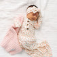 A newborn baby wearing the Lou Lou & Company Baby Girls’ Lottie Knotted Gown with a matching headband rests on a white quilt, gently holding a textured pink blanket, eyes closed—the gown’s convenient button-down front makes diaper changes effortless.