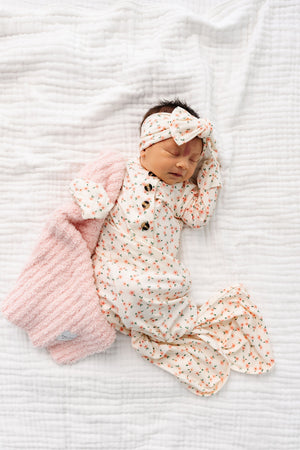 A newborn baby wearing the Lou Lou & Company Baby Girls’ Lottie Knotted Gown with a matching headband rests on a white quilt, gently holding a textured pink blanket, eyes closed—the gown’s convenient button-down front makes diaper changes effortless.