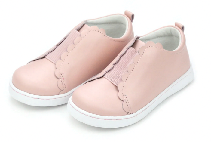 The Girls' Phoebe Slip On Sneaker by L'Amour Shoes features light pink scalloped detailing, a comfy memory foam leather sockbed, and white soles, shown on a white background.