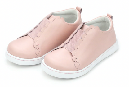 The Girls' Phoebe Slip On Sneaker by L'Amour Shoes features light pink scalloped detailing, a comfy memory foam leather sockbed, and white soles, shown on a white background.
