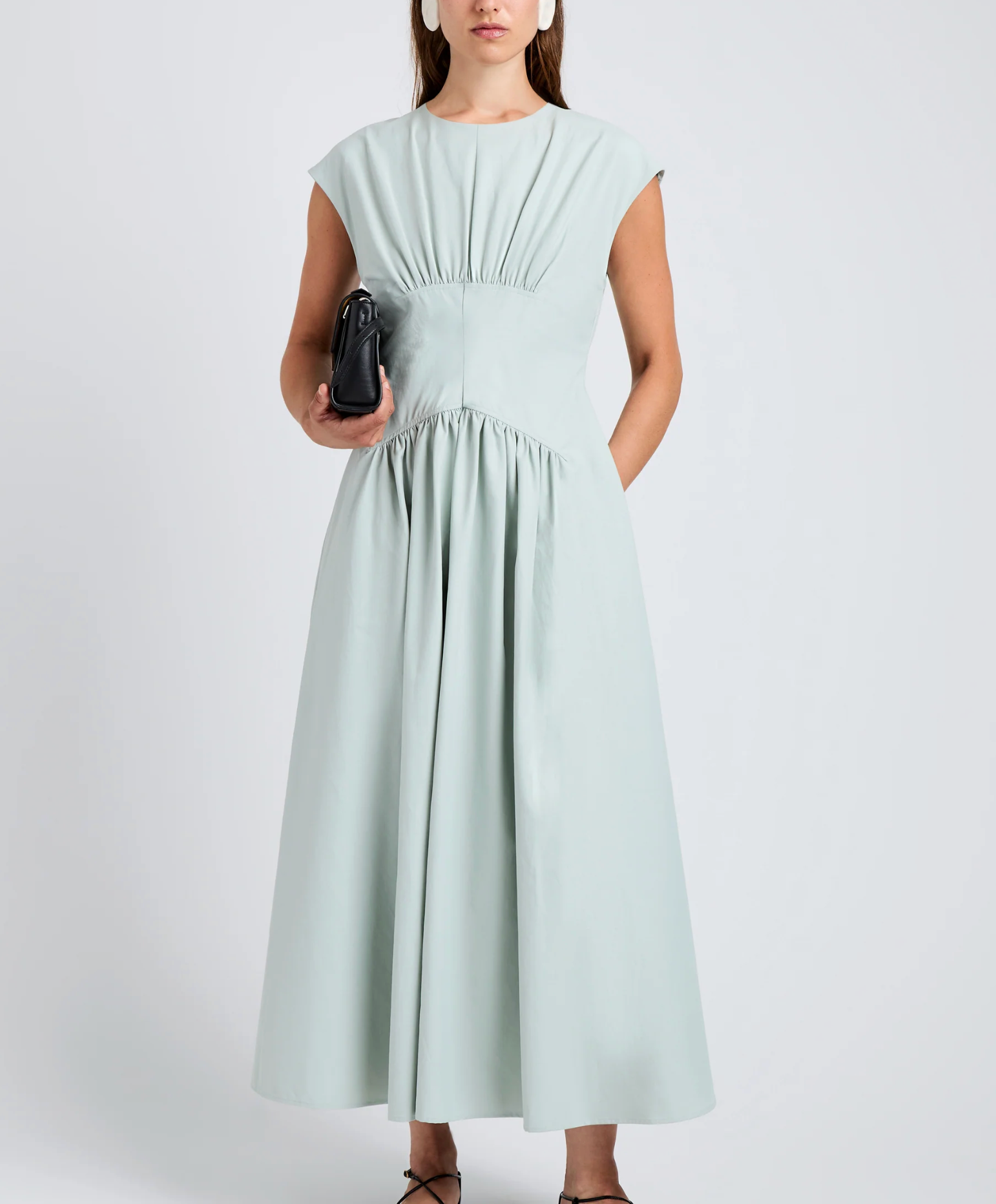 A woman in the Nyssa Dress by Proenza Schouler White Label—a light blue, sleeveless A-line piece with delicate shirring—holds a black clutch against a plain background.