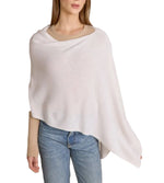 A woman wearing a beige sweater, light blue jeans, and the Alashan Cashmere Dress Topper Poncho in white stands against a white background, effortlessly exemplifying the versatile elegance of this staple piece—perfect for transitioning seamlessly from day to night.