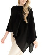 A woman with loose wavy hair stands against a white background, wearing the Alashan Cashmere Cashmere Dress Topper Poncho in black, styled as a versatile wardrobe staple over light-colored pants.