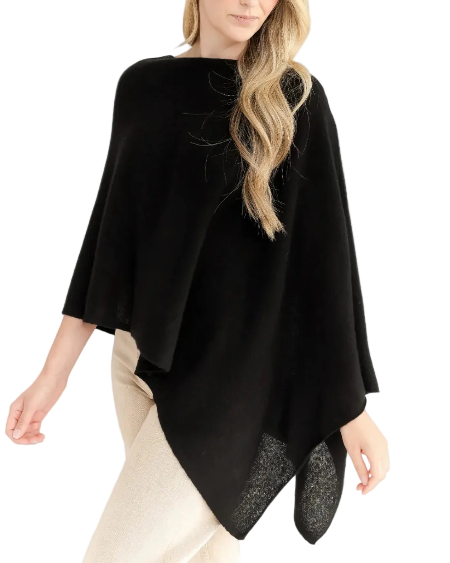 A woman with loose wavy hair stands against a white background, wearing the Alashan Cashmere Cashmere Dress Topper Poncho in black, styled as a versatile wardrobe staple over light-colored pants.
