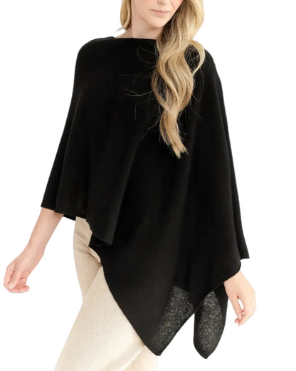 A woman with loose wavy hair stands against a white background, wearing the Alashan Cashmere Cashmere Dress Topper Poncho in black, styled as a versatile wardrobe staple over light-colored pants.