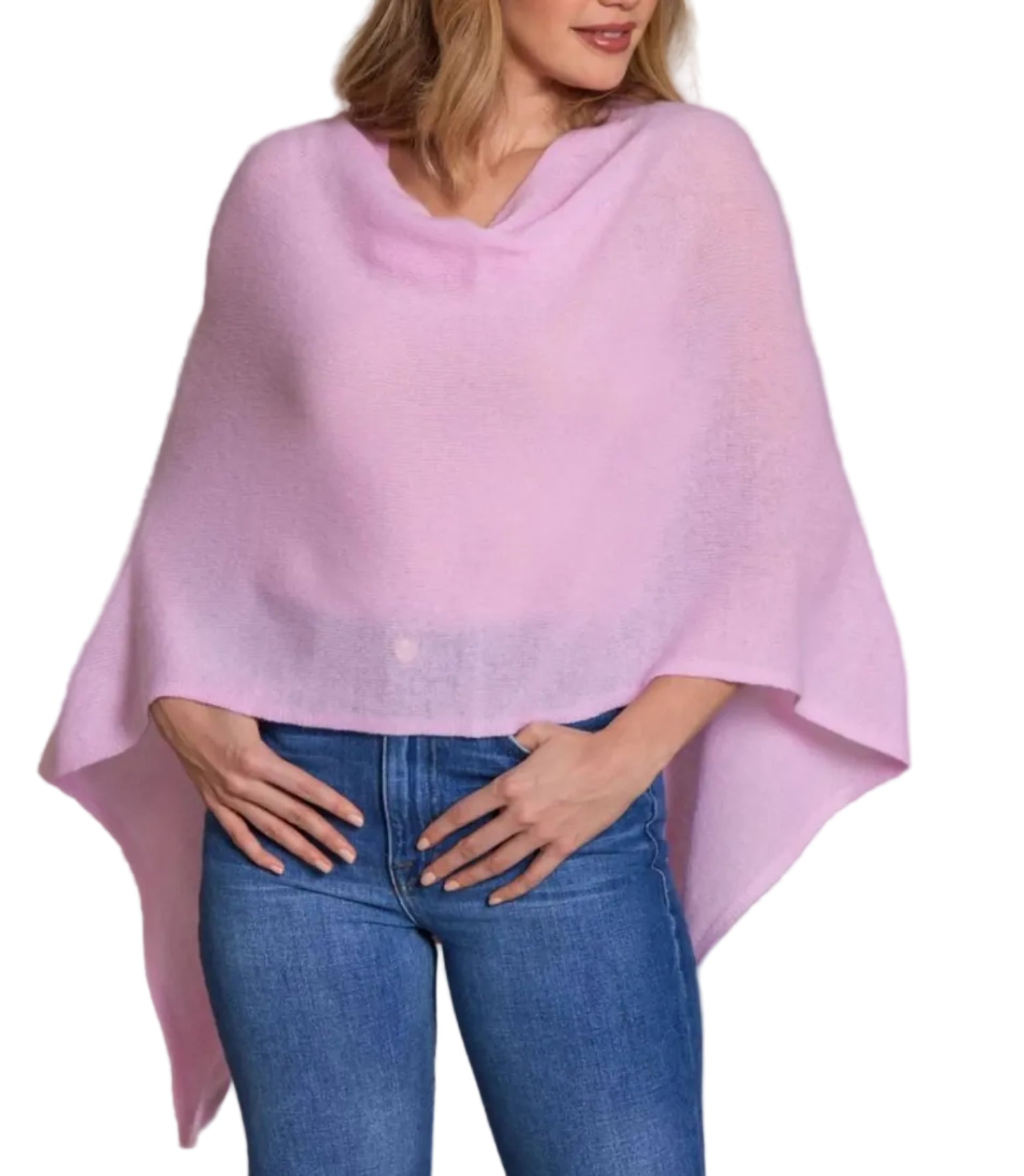 A woman models the Alashan Cashmere Cashmere Dress Topper Poncho in light pink—an effortless wardrobe essential—paired with blue jeans and her hands resting on her hips.