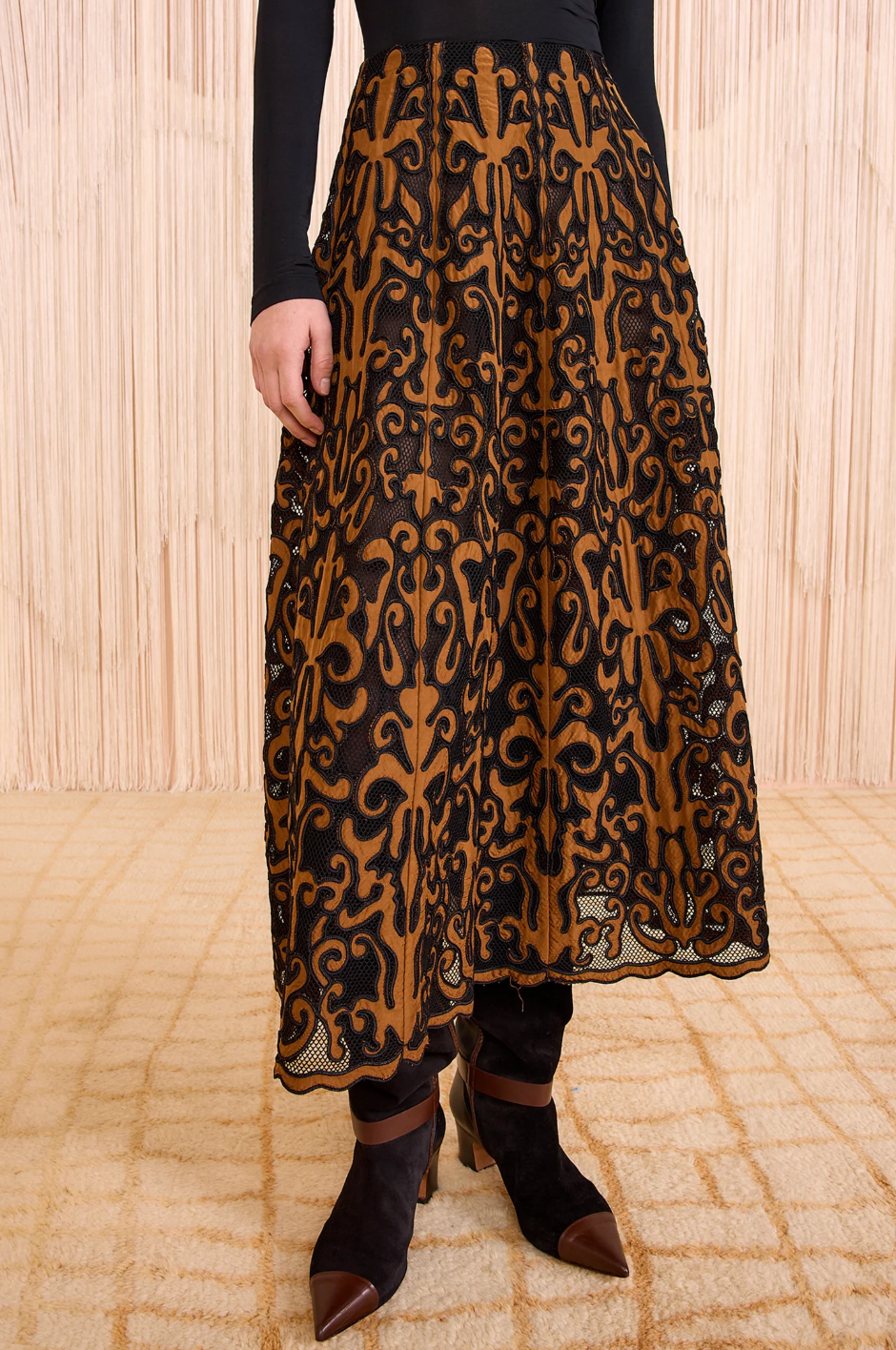 Person wearing a black long-sleeve top, Ulla Johnson's Gili Midi Skirt with brown and black patterns, black leggings, and black pointed-toe ankle boots on a patterned floor.