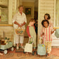 An elderly woman stands on a patio outside a house, her three children eagerly clutching toddler-size baskets and carts filled with toys. Among them, an Olli Ella Rattan Original Luggy adds a charming touch to the scene.