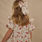 A blonde girl stands facing away, wearing the Girls' Cordelia Dress by Rylee & Cru—a floral ruffle dress with elasticized sleeves and a matching large bow in her hair against a neutral background.