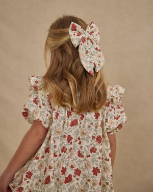 A blonde girl stands facing away, wearing the Girls' Cordelia Dress by Rylee & Cru—a floral ruffle dress with elasticized sleeves and a matching large bow in her hair against a neutral background.