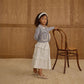 A young girl wears the Rylee & Cru Girls' Ruffled Midi Skirt with tiered layers and an elastic waistband, standing by a wooden chair indoors against a backdrop of wood paneling and carpeted flooring, looking at the camera.