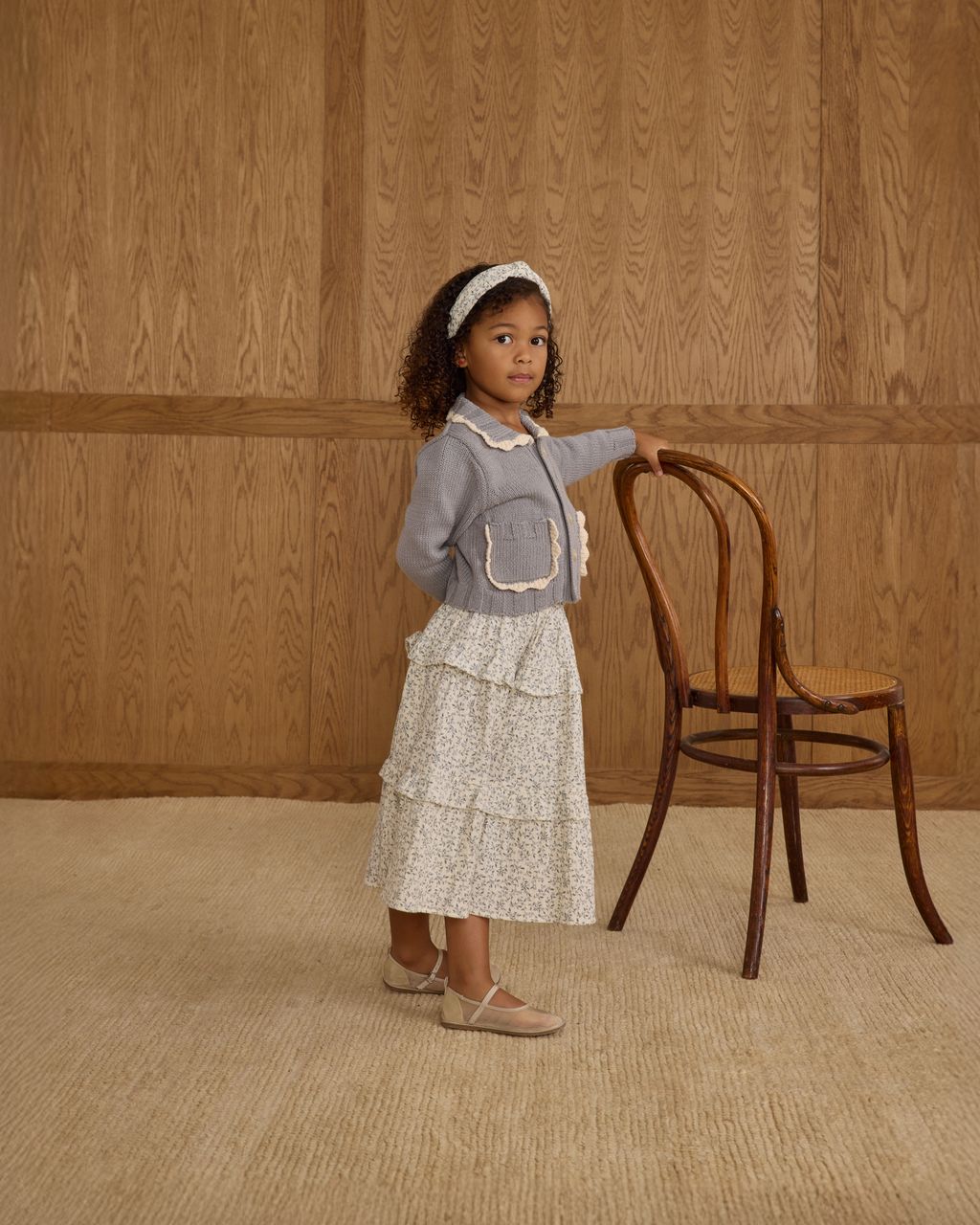 A young girl wears the Rylee & Cru Girls' Ruffled Midi Skirt with tiered layers and an elastic waistband, standing by a wooden chair indoors against a backdrop of wood paneling and carpeted flooring, looking at the camera.