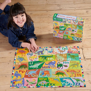 A young girl smiles while lying on a wooden floor, assembling the Eeboo Alphabet of Dinosaurs 36-Piece Giant Puzzle—a vibrant educational toy for children—with its box beside her.