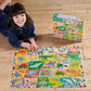 A child smiles on a wooden floor while assembling the colorful Eeboo Alphabet of Dinosaurs 36 Piece Giant Puzzle, with the puzzle box nearby.