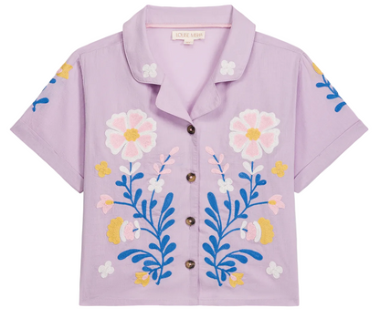 The Girls’ Alova Shirt by Louise Misha is a charming short-sleeve lilac blouse crafted from cotton veil, featuring a classic collar, brown buttons, and colorful floral embroidery on the front—a delightful addition to your wardrobe.
