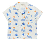 The Arsene & Les Pipelettes Boys' Magnum Shirt is a short-sleeve white double voile shirt featuring blue wave and plant motifs, orange sun accents, a chest pocket, and brown buttons, showcased against a plain background.