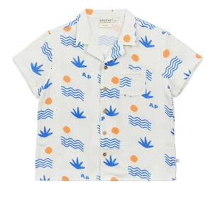 The Arsene & Les Pipelettes Boys' Magnum Shirt is a short-sleeve white double voile shirt featuring blue wave and plant motifs, orange sun accents, a chest pocket, and brown buttons, showcased against a plain background.